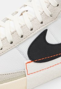 Baskets blanches avec des accents en suède gris clair, présentant un logo noir et des coutures oranges. Matériau texturé et lacets plats.