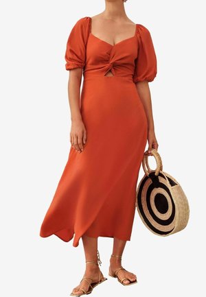 Mujer con vestido midi naranja óxido con mangas abullonadas y nudo delantero, sosteniendo un bolso redondo tejido con un patrón circular negro.