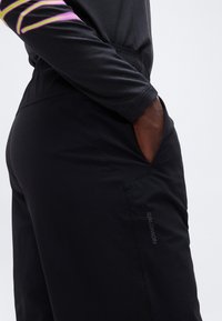 Άτομο που φοράει μαύρο παντελόνι Salomon με το χέρι στην τσέπη και μαύρο μακρυμάνικο τοπ με μωβ και κίτρινες ρίγες.