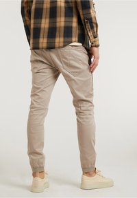 Ljusbeiga tapered byxor med elastiska muddar, två bakfickor och en mjuk textur, ihop med en brun rutig skjorta och beiga sneakers.