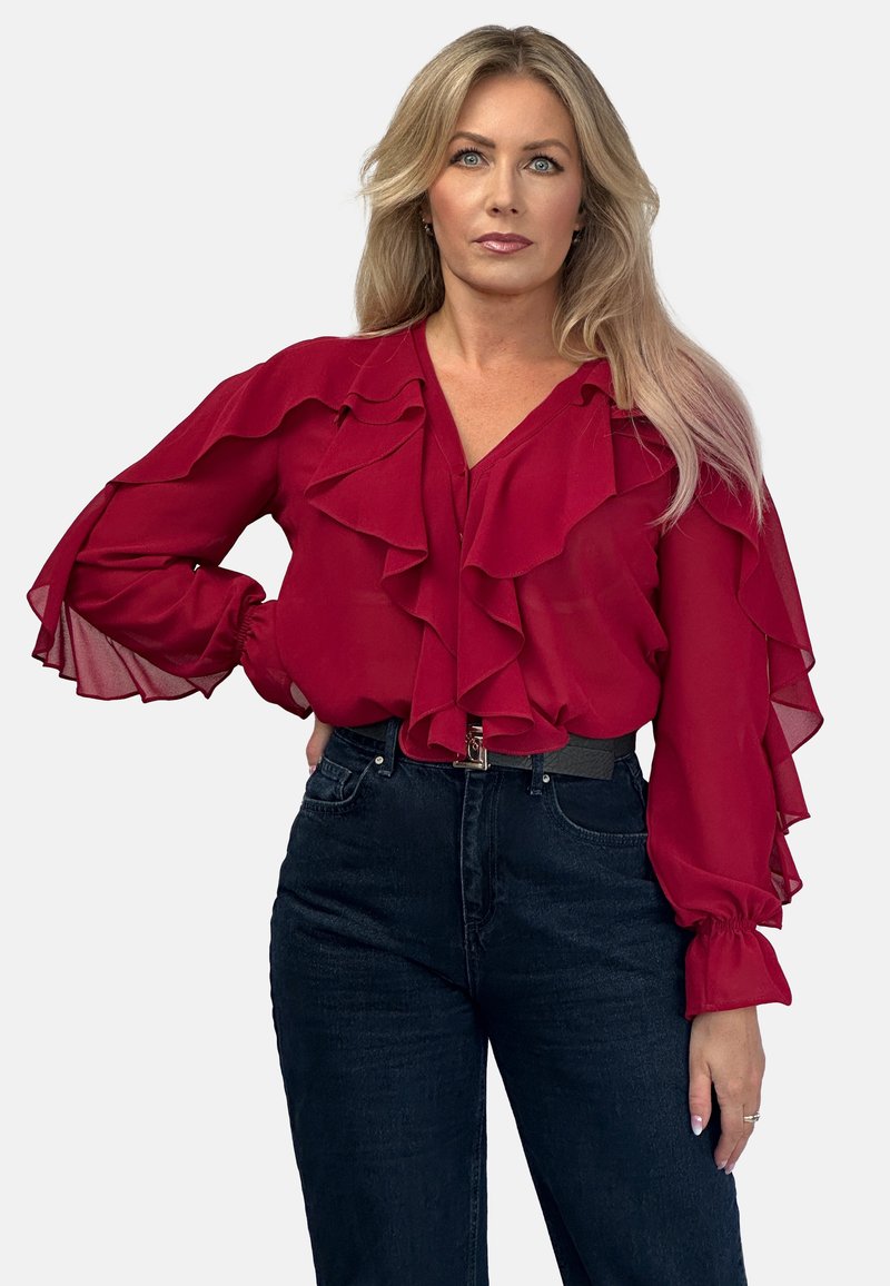 Czerwona bluzka z szyfonu z falbaniastym przodem i rękawami, połączona z ciemnymi dżinsami. Bluzka ma dekolt w kształcie litery V i elastyczne mankiety.