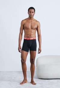 Fekete szűk fazonú boxeralsó piros Nike derékpánttal. A modell mezítláb áll, bemutatva a boxeralsót és a sima textúrát. A háttér semleges.