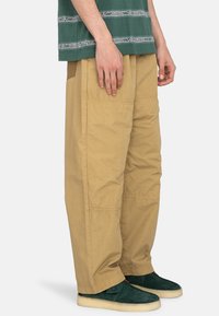 Pantalons cargo beiges avec une coupe décontractée, fabriqués en tissu résistant, dotés de poches latérales et de coutures visibles. Portés avec des chaussures vertes.