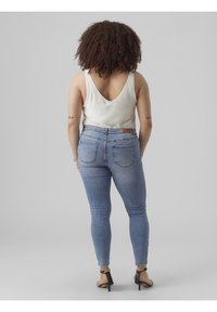 Žena s kudrnatými vlasy, oblečená v bílém topu bez rukávů, světlomodrých skinny džínách a černých sandálech na podpatku, stojící zády k šedému pozadí.