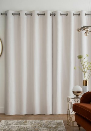 Floor-length beige curtains hanging on a rod in a living room with a brown sofa, patterned rug, side table, and decorative plants.