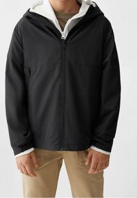 Svart zip-up jacka med huva och vit foder. Har elastiska ärmslut och två framfickor. Slät textur, lätt design.