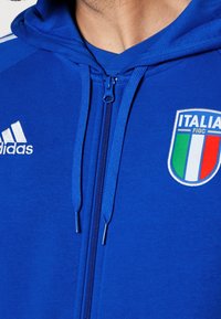 Sweat à zip bleu en tissu doux avec capuche réglable, logo Adidas blanc et emblème de l'équipe nationale d'Italie avec des accents tricolores.