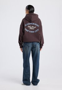 Sudadera marrón con texto azul y blanco en la parte de atrás que dice "BUENOS DÍAS NUEVA YORK HOTEL NUEVA YORK CITY." Combinada con unos jeans oscuros y holgados.