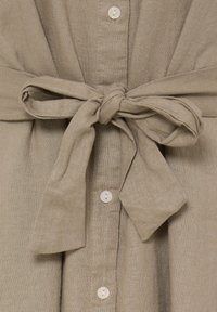 Beige hørstof bundet i en sløjfe i taljen med synlige hvide knapper, der løber lodret under knuden.