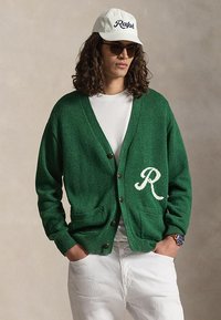 Mężczyzna w zielonym, zapinanym na guziki kardiganie z białą literą "R", białej koszulce, białej czapce z napisem "Ralph", okularach przeciwsłonecznych, białych spodniach i zegarku na rękę na jednolitym tle.