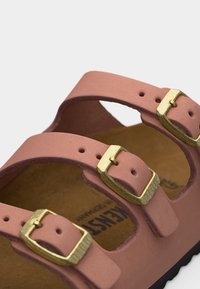 Ροζ σανδάλι με δύο ρυθμιζόμενες χρυσές αγκράφες και καφέ επένδυση στο πέλμα, τοποθετημένο πάνω σε απλό ανοιχτόχρωμο φόντο.