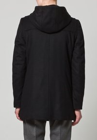 Cappotto lungo in lana nera con un grande cappuccio, caratterizzato da una forma sartoriale, cuciture verticali e una superficie liscia e opaca.