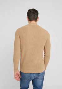 Beige gerippter Pullover mit rundem Halsausschnitt, langen Ärmeln und einem enganliegenden Bündchen, kombiniert mit blauen Denim-Jeans.