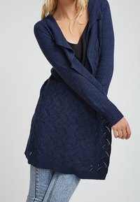 Navy blue knitted cardigan with a wave pattern, open front, long sleeves, and a relaxed fit, featuring textured detailing and side slits.