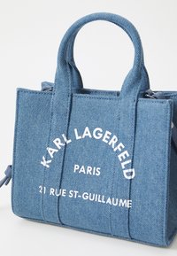 Sac en denim avec double poignées, présentant un texte imprimé en blanc : "KARL LAGERFELD PARIS 21 RUE ST-GUILLAUME." Forme structurée, couleur bleue.