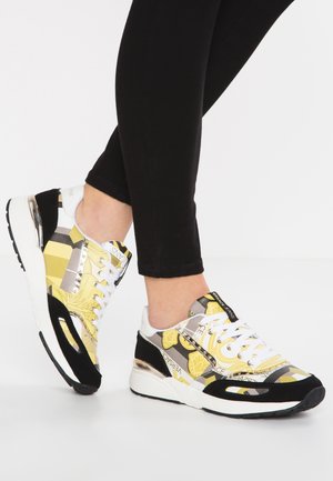 Pies con leggings negros y zapatillas deportivas estampadas en amarillo, negro y blanco con cordones blancos y suela blanca gruesa.