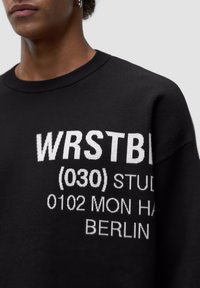 Svart sweatshirt med rund halsringning och lös passform, prydd med vit grafisk text som inkluderar "WRSTB" och "STUDIOS 0102 MON HAF BERLIN."