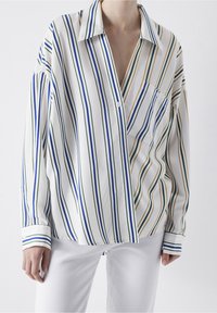 Chemise à manches longues rayée blanche avec des rayures verticales vertes, bleues et jaunes. Comprend une petite poche sur la poitrine et un col.
