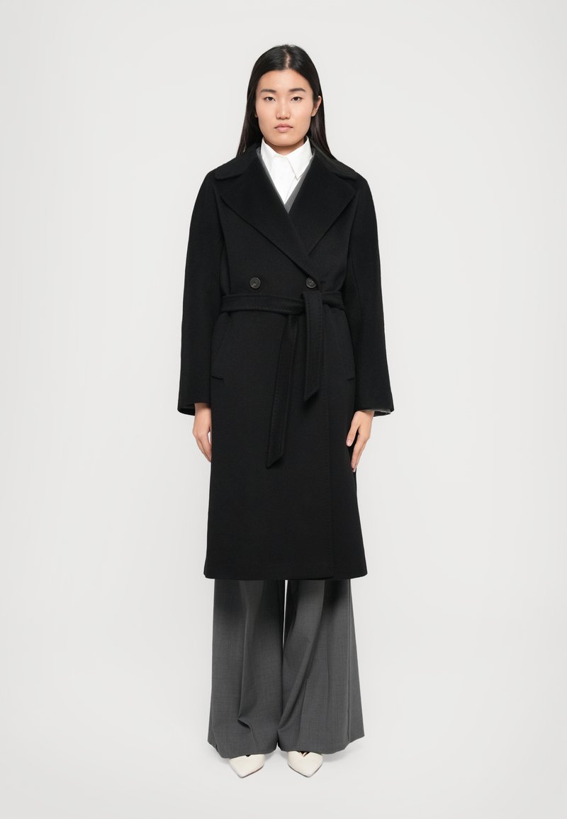 Double-breasted black wool coat with a belted waist, wide lapels, and side pockets, worn over wide gray trousers and pointed white shoes.