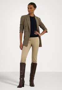 Brown plaid double-breasted blazer, navy top, beige fitted pants, and tall brown boots with a chunky heel. Smooth fabric textures and slim fit.
