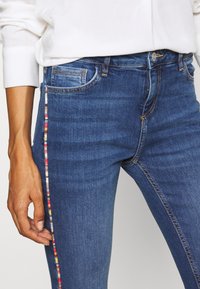Los jeans de mezclilla azul ajustados presentan una franja lateral con detalles tejidos en múltiples colores y un diseño estándar de cinco bolsillos. Con costuras sutiles que los destacan.