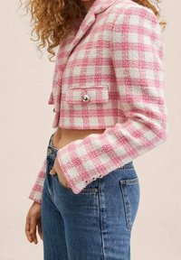 Zkrácená bunda v růžovém a bílém kostkovaném vzoru, z texturované tkaniny, se dvěma předními kapsami a stříbrnými knoflíkovými detaily, nošena přes džíny.