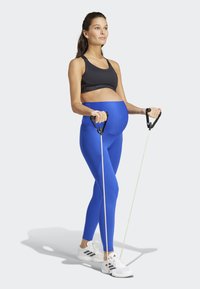 Top sportivo nero, leggings azzurri a vita alta, scarpe da ginnastica bianche. Tiene in mano bande di resistenza con maniglie nere, all'interno contro uno sfondo bianco.