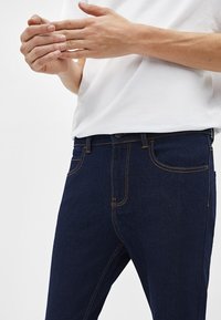 Mörkblå denimjeans med orange sömmar, knappgylf och sidofickor. Bärs med en vit t-shirt, visar en närbild av händer och midja.
