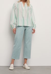 Camicia leggera a righe in bianco, verde e blu, con scollatura a V e maniche lunghe a sbuffo, abbinata a pantaloni a gamba larga verde menta e sneakers bianche.