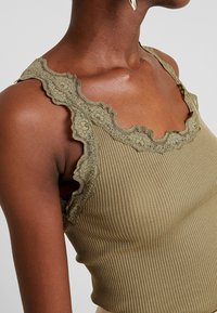 Close-up of a person wearing a ribbed olive green tank top with floral lace trim on straps and neckline, paired with a gold earring.