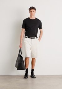 Schwarzes T-Shirt, weiße Shorts, schwarzer Gürtel, schwarze Socken und schwarze Schuhe. Hält eine große schwarze Tragetasche mit strukturiertem Material.