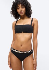 Γυναίκα με σγουρά μαλλιά που φοράει μαύρο ριχτό μπικίνι τοπ με χρυσό κούμπωμα και μαύρο μπικίνι κάτω μέρος με λευκή, ταμπά και μαύρη πλεξούδα στο τελείωμα.