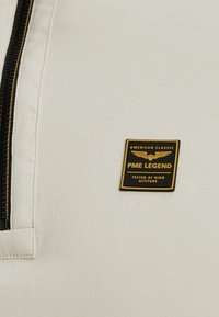 Tela de sudadera blanca con una cremallera negra y una etiqueta cuadrada en negro y dorado que dice "PME LEGEND AMERICAN CLASSIC TESTED AT HIGH ALTITUDE."