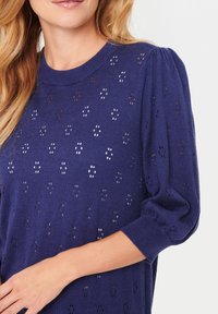 Marinblå kortärmad tröja med puffärm, med ett texturerat mönster av små utskärningar, rund halsringning och ribbad mudd.