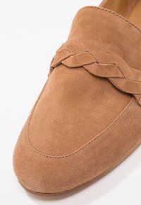 Mocassin en daim marron clair avec bout pointu et détail de bride tressée sur le dessus.