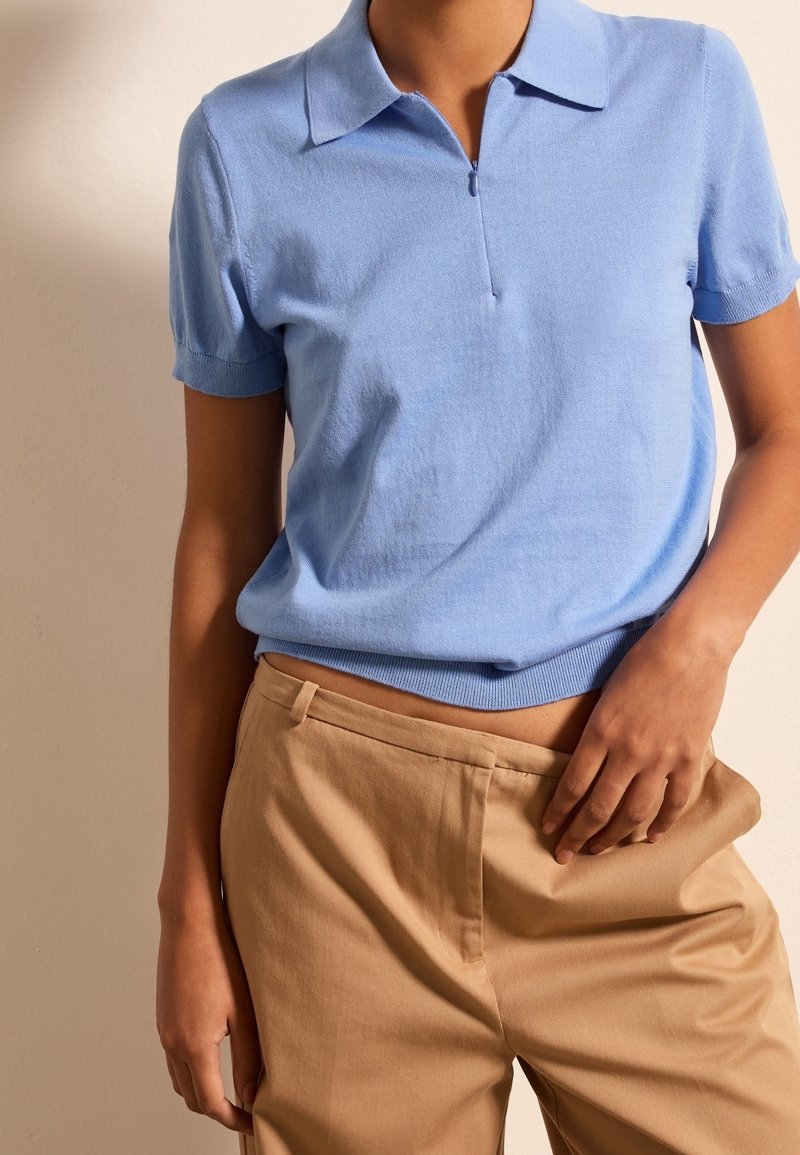 Personne portant une chemise bleu clair à manches courtes et col, rentrée dans un pantalon beige taille haute, avec une main posée sur la hanche.