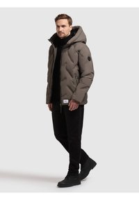 Giacca Puffer verde oliva, con finitura lucida, cappuccio oversize, cuciture diagonali e un'etichetta logo nera sul braccio sinistro.