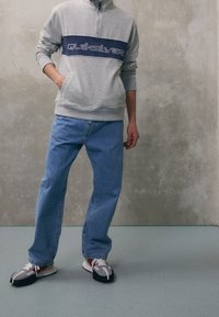 Persona vistiendo un sudadera gris de Quiksilver, jeans azules holgados y zapatillas grises con calcetines rojos, de pie contra una pared texturizada.