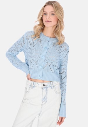 Donna che indossa un maglione corto azzurro a bottoni realizzato con maglia aperta e jeans a vita alta chiari, posando con una mano sul fianco.