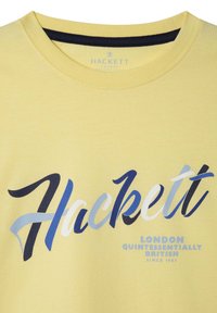 Żółta bawełniana koszulka z okrągłym dekoltem z wyraźnym logo "Hackett" w niebieskim i czarnym, z napisem "London Quintessentially British" poniżej.