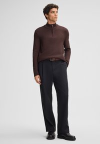 Brauner, strukturierter Halbzip-Pullover kombiniert mit schwarzen, gefalteten Hosen und schwarzen Schuhen. Das Ensemble hat eine lässige Passform und einen Gürtel.