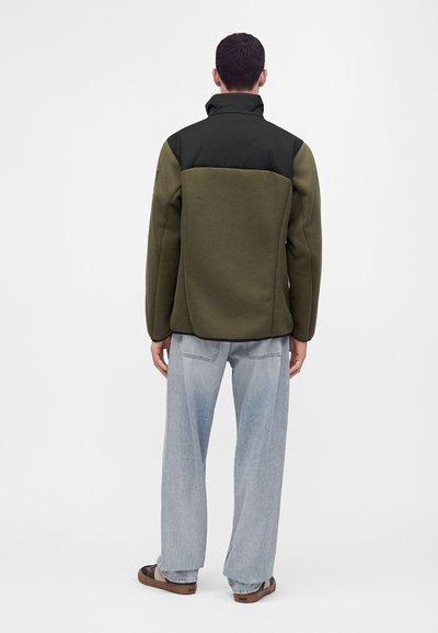Homme portant une veste vert olive et noire, un jean large bleu clair, et des sandales marron, debout de dos devant un fond uni.