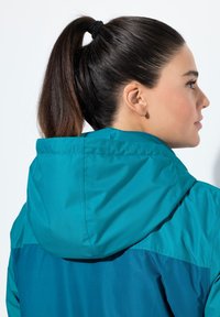 Grönblå vindjacka med huva, med en jämn textur och färgblockering vid axlarna. Designen är strömlinjeformad.