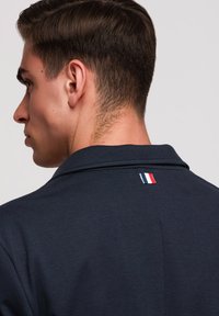 Junger Mann mit kurzen braunen Haaren trägt ein dunkelblaues Hemd mit einem kleinen rot-weiß-blau gestreiften Etikett am hinteren Kragen.