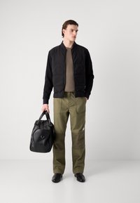 Veste bomber noire en tissu matelassé, pull en tricot marron, pantalon large vert olive, et sac en cuir noir. Matériaux lisses, détails matériels minimaux.