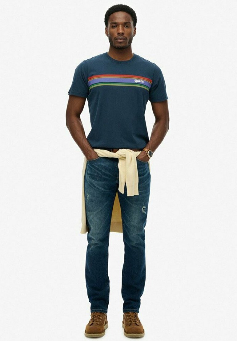 Camiseta de algodón azul marino con rayas horizontales multicolores, combinada con jeans desgastados de color azul claro y botas de cordones marrones. Suéter atado a la cintura.