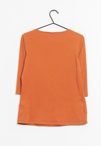 Orange bomullstopp med trekvartslånga ärmar, rund hals och rak skärning. Tyget har en mjuk textur och hänger från en svart krok.