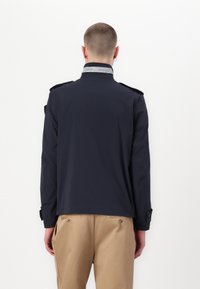 Marineblauwe jas met een gestructureerde kraag en schouderepauletten, voorzien van een zilveren logo-accent bij de nek, gemaakt van lichtgewicht materiaal.