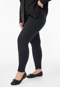 Leggings negras de un tejido texturizado, diseño ajustado, combinados con bailarinas negras con un lazo decorativo. Fondo neutro.