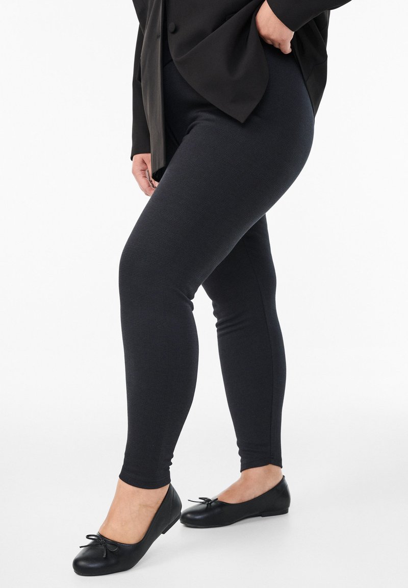 Leggings negras de un tejido texturizado, diseño ajustado, combinados con bailarinas negras con un lazo decorativo. Fondo neutro.